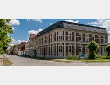 Ecke einer gepflasterten Straße mit einem historischen, zweigeschossigen Gebäude aus rotem Backstein und weißen Fenstereinfassungen. Im Vordergrund stehen vereinzelt kleine Bäume, und der Hintergrund zeigt weitere Backsteinhäuser unter blauem Himmel mit einigen Wolken.