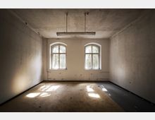 Ein leerer Raum mit zwei großen Fenstern mit Rundbögen, durch die Tageslicht einfällt. Der Raum hat abgenutzte Wände, einen teilweise beschädigten Boden und eine Hängelampe an der Decke.