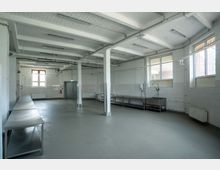 Großer, leerer, industriell anmutender Raum mit weiß gestrichenen Wänden, freiliegenden Deckenbalken und großen Fenstern. Der Raum ist mit Edelstahl-Arbeitsflächen und einem Edelstahlspülbecken ausgestattet; Fliesen und Fußboden sind in hellem Grau gehalten.