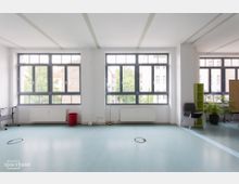 Heller Raum mit großen, rechteckigen Fenstern, die Blick auf umliegende Gebäude und Bäume bieten. Der Raum hat einen blauen Bodenbelag, weiße Wände, Heizkörper unter den Fenstern und einige Möbel wie einen Stuhlstapel, ein Regal und Pflanzen.