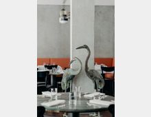 Innenraum eines modernen Restaurants mit einer hellgrauen Betonwand und einer quadratischen Säule, an der zwei bronzene Kranichstatuen lehnen. Im Vordergrund sind gedeckte Tische mit Glasplatten, weißen Servietten und Gläsern zu sehen, im Hintergrund gibt es orangene Sitzbänke und schwarze Stühle.