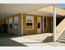 Ein flacher überdachter Gebäudebereich mit gelblichen Backsteinwänden, Schaufenstern mit der Beschriftung „Bade- und Strandartikel“ sowie „Bademode Süßwaren“ und einer Eingangstür. Rechts führt eine Außentreppe nach oben, und links ist eine Tafel mit einem Essensangebot aufgestellt.