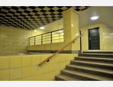 Unterirdischer Bereich mit gelben gefliesten Wänden und einer Decke mit schwarzem-weißem Schachbrettmuster. Eine Treppe führt zu einer erhöhten Plattform mit Geländer und einer schwarzen Tür.