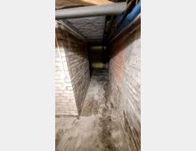 Ein schmaler Kellerflur mit Wänden aus unverputzten Ziegelsteinen und einem Betonboden, der feuchte Stellen aufweist. An der Decke verlaufen Rohre und Kabel, und am Ende des Flurs ist eine gelbliche Wand erkennbar.