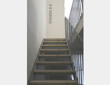 Treppe mit offenen Holzstufen und Metallgeländern in einem minimalistischen Innenraum; an der Wand oben links ist eine vertikale Beschriftung zu sehen.