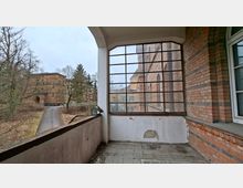 Überdachter Laubengang oder Balkon an einem Backsteingebäude mit Fliesenboden und einer großen, gitterartig gerahmten Fensterfront. Links führt ein Weg durch kahlen Baumbestand zu weiteren Backsteinbauten im Hof.