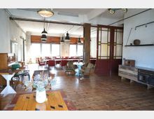 Industrie-inspirierter Innenraum mit Holzboden, großen Fenstern, roten Stühlen und Pendelleuchten. Im Vordergrund stehen ein Tisch mit Blumen in einer Vase sowie weitere Sitzmöbel, und eine dekorative Holztür mit Glaselementen trennt den Raum.