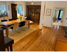 Esszimmer und Wohnzimmer in einem hellen, modernen Zuhause mit Holzdielenboden. Ein großer Esstisch mit acht Stühlen steht auf einem Teppich, daneben ein Ledersofa; im Hintergrund sind weiße Wände, ein antiker Holzschrank und Fenster mit Vorhängen zu sehen.