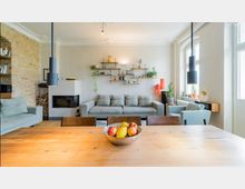 Modernes Wohnzimmer mit einer hellgrauen Couch, einer Wand mit offenen Regalen, auf denen Pflanzen und Dekorationen stehen, sowie einem Kamin in der linken Ecke neben einer Backsteinwand. Im Vordergrund steht ein hölzerner Esstisch mit einem Obstkorb, und große Fenster lassen viel Tageslicht herein.