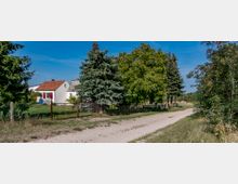 Ländliche Szene mit einem kleinen weißen Haus mit roten Dachziegeln und roten Fensterläden, umgeben von einem Garten mit Bäumen und einem Drahtzaun. Im Vordergrund führt ein unbefestigter Weg an der umzäunten Grundstücksgrenze entlang.