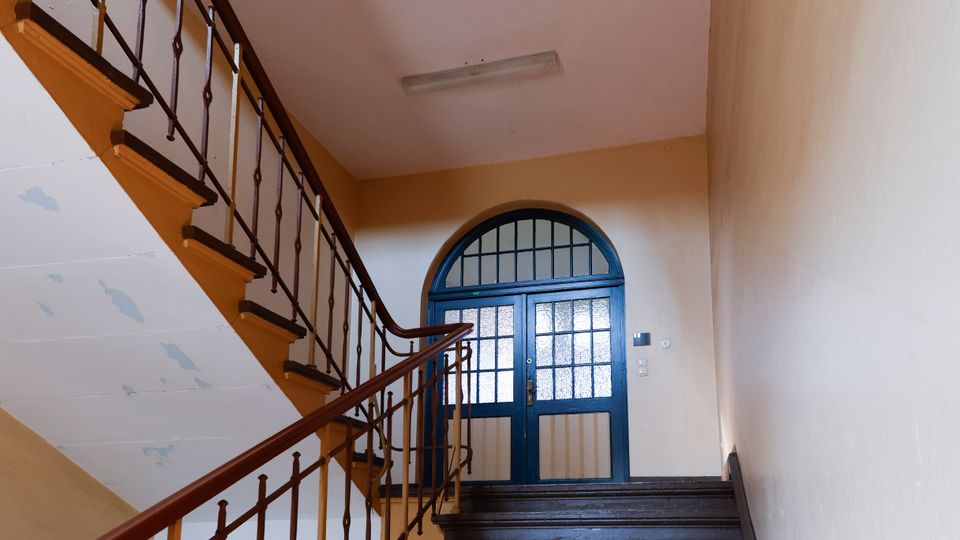 Treppenhaus in einem älteren Wohngebäude mit dunklen Holzstufen und Metallgeländer sowie Holzhandlauf. Am oberen Absatz befindet sich eine blau gestrichene Doppeltür mit halbkreisförmigem Oberlicht und strukturierten Glasfeldern.