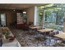 Ein gemütlicher Innenraum mit einer Sitzgruppe aus roten Polstermöbeln und Holztischen mit blauen Stühlen. Eine Wand ist mit Filmplakaten dekoriert, während große Fenster Tageslicht hereinlassen und den Blick auf Bäume im Außenbereich freigeben. Auf der linken Seite steht ein Buffet mit Salaten und Speisen.