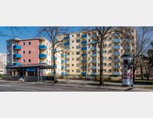 Mehrstöckige Wohngebäude mit hellgelber und rosa Fassade sowie blauen Balkonen, umgeben von kahlen, blattlosen Bäumen. Im Vordergrund eine Straßenlaterne, eine Litfaßsäule mit Werbung und ein Eingang zu einer U-Bahn-Station.