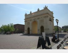 Historisches Stadttor auf einem gepflasterten Platz, flankiert von verzierten Reliefs und Figuren auf der Oberseite. Im Hintergrund stehen Bäume und Gebäude aus rotem Backstein, während im Vordergrund Stühle und eine Straßenlaterne sichtbar sind.