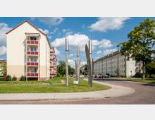 Mehrstöckige Wohngebäude mit beigefarbenen Fassaden und roten Balkonbrüstungen säumen eine Grünfläche mit einer modernen Metallskulptur in der Mitte. Im Hintergrund sind geparkte Autos, ein gepflasterter Gehweg und Bäume zu sehen, unter einem Himmel mit vereinzelten Wolken.