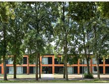 Zweistöckiges Gebäude mit einer Fassade aus orangefarbenem Backstein und großen schwarzen Fenstern, umgeben von hohen Bäumen in einem parkähnlichen Umfeld. Ein Weg führt entlang der Vorderseite des Gebäudes durch die Grünfläche.