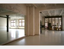 Großer Raum mit weiß gestrichenen Backsteinwänden und einer glänzenden, hellen Bodenfläche, der als Tanz- oder Übungsraum dient. Links befindet sich eine Spiegelwand, rechts sind große Glastüren, die zu einem angrenzenden Bereich mit Möbeln führen.