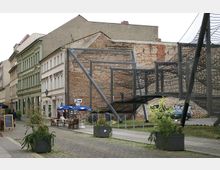 Straßenszene mit historischen Gebäuden, darunter eine grüne Fassade, und einem modernen Klettergerüst aus Netzen und Metallstangen im Vordergrund. Im Außenbereich eines Cafés stehen Tische und Stühle unter blauen Sonnenschirmen, flankiert von Pflanzkübeln.