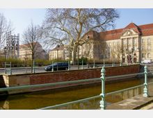 Historisches Gebäude mit heller Fassade und roten Ziegeldächern, umgeben von Bäumen ohne Laub. Im Vordergrund fließt ein schmaler Kanal, gesäumt von einer Backsteinmauer und einem grün gestrichenen Geländer.