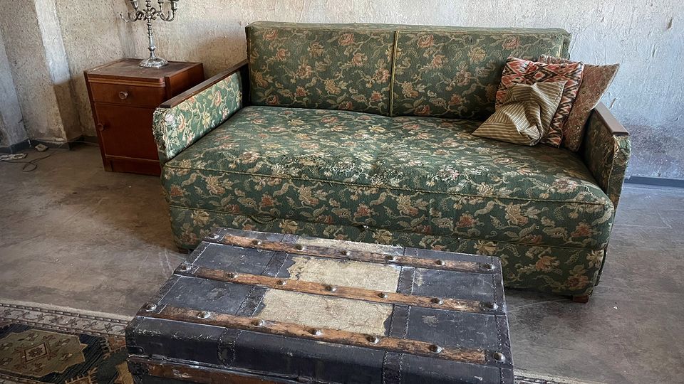Rustikaler Innenraum mit rau verputzten Wänden und Betonboden: Im Mittelpunkt steht ein grünes, gemustertes Sofa mit Kissen, davor eine große schwarze Truhe mit Metallbeschlägen. Links daneben steht ein kleiner Holz-Nachttisch mit silbernem Kerzenleuchter, teils auf einem gemusterten Teppich.