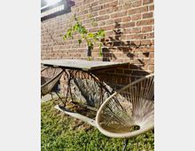 Ein schlichter Außenbereich mit einem Holztisch und zwei runden Stühlen mit geflochtenen Sitzflächen, vor einer Ziegelsteinwand. Auf dem Tisch steht eine Glasvase mit grünen Zweigen, und der Boden besteht aus Rasen.