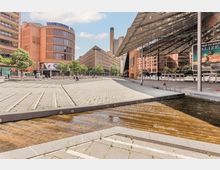 Offener Platz mit modernem urbanem Design, umgeben von Gebäuden mit Glasfassaden und Backsteinarchitektur. Im Vordergrund ein Wasserlauf mit Treppenstruktur, im Hintergrund das Theatergebäude "Blue Man Group" und weitere Geschäftsgebäude.