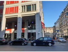 Straßenszene mit einem modernen Gebäude, das großflächige Fenster und rote Fassadenelemente aufweist, darunter ein Ladengeschäft mit Beschriftungen wie "Film" und "Musik-Noten". Davor parken Autos, und im Hintergrund sind weitere Gebäude sowie der blaue Himmel sichtbar.