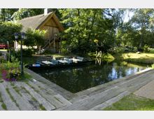 Holzhaus mit Reetdach an einem ruhigen, schmalen Kanal, umgeben von Bäumen und Blumen. Im Vordergrund ein Steg sowie ein Boot mit Sitzplätzen; dahinter Holzbrücken und eine Wiese im Gartenbereich.