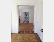 Blick durch eine offene Tür in einen hellen Wohnraum mit Parkettboden und weißen Wänden. Im Hintergrund ein Esstisch mit weißen Stühlen vor einem großen Fenster, das Tageslicht hereinscheinen lässt.