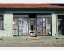 Fassade eines alten Gebäudes mit grüner, abblätternder Farbe. Zu sehen sind eine Tür und große Fenster mit Gitterstäben, hinter denen Gardinen und verschiedene Gegenstände erkennbar sind; davor führt eine kleine Stufe vom gepflasterten Gehweg hinauf.