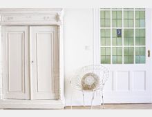 Ein heller Innenraum mit einem weißen, antiken Schrank links, einer Tür mit grünem Glas und weißen Holzrahmen rechts, sowie einem weißen Stuhl mit einem dekorativen, floralen Kissen im Vordergrund. Der Boden besteht aus hellem Holzparkett.