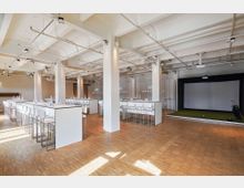 Moderner Veranstaltungsraum mit hellem Parkettboden, weißen Wänden und hohen Decken mit freiliegenden Rohren und Beleuchtung. Der Raum ist mit hohen weißen Tischen und Barhockern möbliert, rechts befindet sich ein Golf-Übungsbereich mit einer großen Leinwand und Kunstrasen.