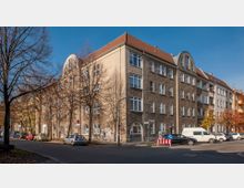 Eckgebäude eines mehrstöckigen Wohnhauses mit hellem Putz, großen Fenstern und einem roten Satteldach, umgeben von kahlen Bäumen in einer ruhigen Wohnstraße. Einige parkende Autos und ein Gehweg mit Straßenschildern sind sichtbar.