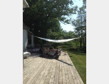 Eine Holzterrasse im Außenbereich mit einem langen Tisch, an dem mehrere Personen essen und sich unterhalten. Im Hintergrund befinden sich Bäume und eine grüne Wiese, während ein Sonnensegel Schatten auf die Szenerie wirft.