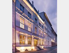 Fassadenansicht eines historischen Gebäudes bei Dämmerung, mit verzierten Fenstergittern, beleuchteten Fenstern und einer dekorativen Fassade mit Reliefs. Das Gebäude steht an einer gepflasterten Straße, flankiert von ähnlichen Reihenhäusern.