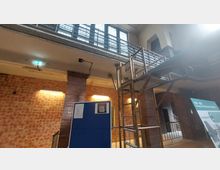 Industriehalle mit Backsteinwänden und einer Metalltreppe, die zu einem oberen Gang mit Geländer führt. Im Vordergrund steht eine blaue Informationswand und ein Baugerüst; im Hintergrund sind geflieste Wände und beleuchtete Notausgangsschilder zu sehen.