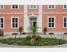 Fassade eines rosafarbenen Gebäudes mit weißen Fensterrahmen und einem ornamentierten weißen Eingangstor in der Mitte. Vor dem Gebäude befindet sich ein runder, bepflanzter Vorgarten mit Blumen, Gebüsch und einer Palme in einem Topf.