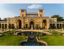 Neoklassizistisches Gebäude mit hellbeiger Fassade, charakterisiert durch Rundbögen, Säulen und zwei rechteckige Türme; davor ein angelegter Garten mit einer Statue in der Mitte und einem symmetrischen Wasserbecken im Vordergrund. Im Hintergrund blauer Himmel mit wenigen Wolken.