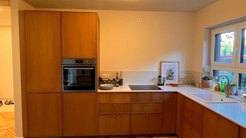 Moderne Küche mit Holzschränken, eingebautem Backofen und weißer Arbeitsplatte; links ein Kochfeld, rechts ein Spülbecken unter einem Fenster. Am Boden mehrfarbige geometrische Fliesen und an der Wand ein sanfter Cremefarbton.