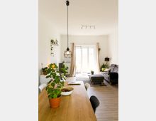 Helle Wohnzimmereinrichtung mit Essbereich im Vordergrund; ein Holztisch mit Topfpflanze und Schalen steht zentral, im Hintergrund ein Sofa, ein Teppich und große Fenster mit Zugang zu einem Balkon. Moderne Hängelampe und minimalistische Dekoration ergänzen die schlichte Gestaltung.
