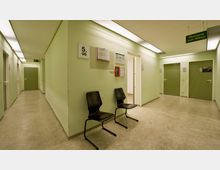 Flur in einem öffentlichen Gebäude mit grünen Wänden, Linoleumboden und einer Deckenbeleuchtung. Der Bereich zeigt grüne Türen, schwarze Stühle an einer Wand, Wegweiser mit der Aufschrift „Übergang zum Altbau“ sowie eine Beschilderung für das 5. Obergeschoss und einen Feuerlöscher.