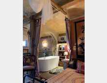 Ein wohnlich gestaltetes Badezimmer mit warmen, erdigen Farbtönen und orientalischen Dekorationselementen. Im Vordergrund steht eine freistehende weiße Badewanne vor einer gemauerten Nische mit indirekter Beleuchtung, umgeben von Holzbalken, bunten Textilien und einem rustikalen Holzfußboden.