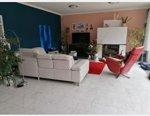 Ein modernes Wohnzimmer mit hellen Marmorböden, einer weißen Couch, einem roten Sessel und einem Kamin. Die Wand ist zweifarbig in Dunkelblau und Rosa gestaltet, mit dekorativen Bildern und Pflanzen, sowie einem großen Fernseher auf einem TV-Schrank.