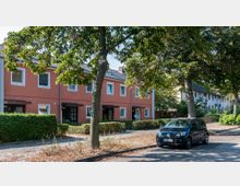 Reihenhäuser mit roten Fassaden und weißen Fenstern, umgeben von kleinen Vorgärten mit niedrigen Hecken. Im Vordergrund verläuft eine Straße mit einem geparkten Auto und schattenspendenden Bäumen.