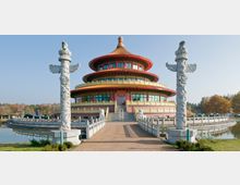 Traditionelle, mehrstöckige chinesische Tempelarchitektur mit runder Form, verziert mit roten und goldenen Details sowie geschwungenem Dach. Im Vordergrund führen symmetrische Treppen und ein gepflasterter Weg über eine Brücke mit zwei reich verzierten weißen Steinsäulen; umgeben von einem kleinen Wassergraben und herbstlicher Landschaft.