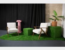 Zwei weiße Sessel mit Holzgestell stehen auf einem Teppich aus künstlichem Gras in einem minimalistisch gestalteten Raum. Im Hintergrund befinden sich ein schwarzer Vorhang, eine rosa Säule und eine Topfpflanze auf einem würfelförmigen Tisch mit Grasoptik.