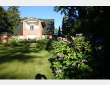 Zweistöckiges Backsteinhaus mit grünem Dach in einem Garten, umgeben von üppiger Vegetation, darunter lila Hortensien und andere Büsche. Im Vordergrund ist eine gepflegte Rasenfläche zu sehen, eingefasst von Schatten und Bäumen im Hintergrund.