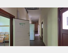 Ein schmaler Flur in einem Gebäude, mit beigen Wänden und mehreren Türöffnungen, darunter eine offene Tür zu einem Klassenzimmer mit Holzstühlen. Über dem Flur befindet sich ein Schild mit der Aufschrift "Gebäude C, 2. OG".