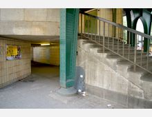 Unterführung aus gelblichen Fliesen mit einem beleuchteten Gang, daneben eine breite Betontreppe mit Metallgeländer. Im Vordergrund steht eine grüne Stahlstütze einer Brückenkonstruktion.