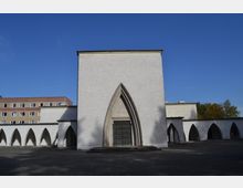 Weiße Kirche mit schlichter Fassade, einem großen Spitzbogenportal und angrenzenden, von Spitzbögen geprägten Arkaden. Im Hintergrund sind ein mehrstöckiges Wohngebäude und herbstliche Bäume sichtbar.